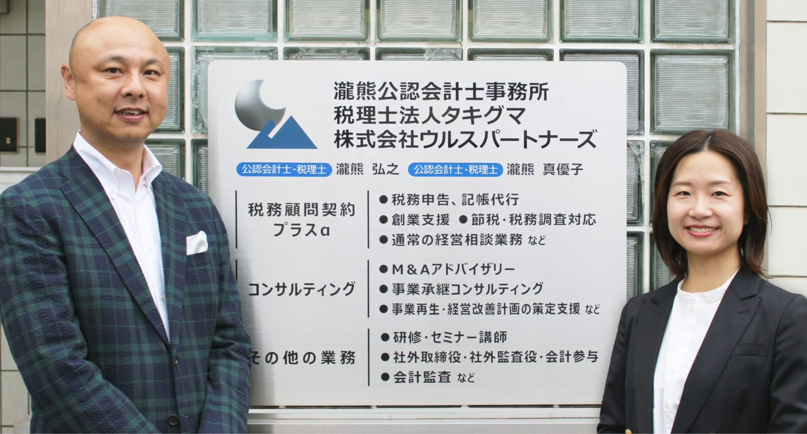 瀧熊弘之と瀧熊真優子の2人が事務所の看板の前で笑顔で立っている様子